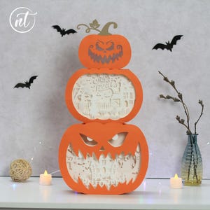 Może przedstawiać: Trzypoziomowa pomarańczowa dekoracja na Halloween z dyni z wyrzeźbionymi twarzami. Środkowa część ma wycięte słowa "Trick or Treat". Czarne wycinanki nietoperzy znajdują się na ścianie za dynią. Mały wazon z gałązkami i świecą stoi na stole.
