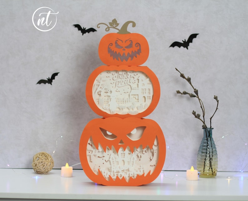 Może przedstawiać: Trzypoziomowa pomarańczowa dekoracja na Halloween z dyni z wyrzeźbionymi twarzami. Środkowa część ma wycięte słowa "Trick or Treat". Czarne wycinanki nietoperzy znajdują się na ścianie za dynią. Mały wazon z gałązkami i świecą stoi na stole.