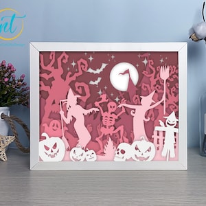 May include: A framed Halloween-themed paper cut design. The artwork features silhouettes of witches, skeletons, pumpkins, and bats in shades of pink and white, set against a full moon backdrop. The frame is white.