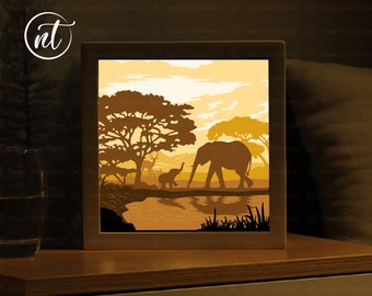 May include: A paper cut-out lamp featuring a silhouette of two elephants walking by a lake in a savanna setting. The lamp is illuminated, casting a warm glow on the scene.