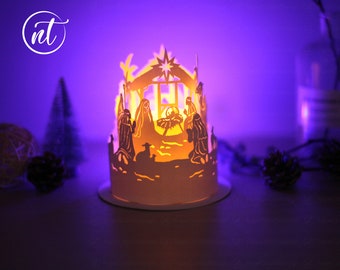 Könnte beinhalten: Eine weiße Papierlaterne mit einem ausgeschnittenen Design der Weihnachtsgeschichte. Die Laterne wird von innen beleuchtet und erzeugt ein warmes Leuchten. Die Szene zeigt Maria, Josef, das Jesuskind, einen Stern und einen Hirten.