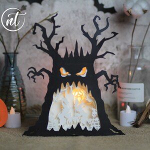 Op de afbeelding: Een zwarte papieren Halloween-decoratie in de vorm van een boom met een griezelig gezicht. De boom heeft een witte binnenkant met een gloeiend licht erin.