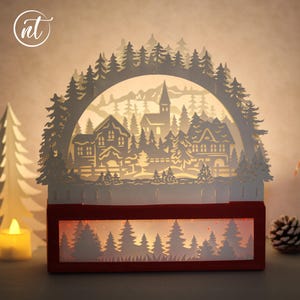 Könnte beinhalten: Ein weißer Papierauschnitt einer Winterdorf-Szene mit Häusern, Bäumen und einer Kirche wird von innen beleuchtet. Die Szene befindet sich in einer roten rechteckigen Box mit einer Waldsilhouette am Boden. Eine Kerze und ein Tannenzapfen sind sichtbar.
