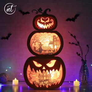 Może przedstawiać: Trzypoziomowa dekoracja z dyni na Halloween z wyrzeźbionymi twarzami i scenami wewnętrznymi. G&oacute;rna dynia ma łodygę i groźny uśmiech. Środkowa dynia m&oacute;wi "Trick or Treat" i pokazuje upiorne miasto. Dolna dynia m&oacute;wi "Happy Halloween".