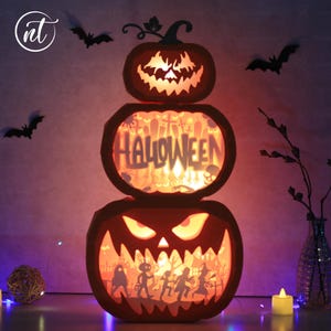 May include: A three-tiered Halloween pumpkin decoration with carved faces and the word "HALLOWEEN". The orange pumpkins are illuminated from within, casting a warm glow. Black bat silhouettes are scattered around the pumpkins.