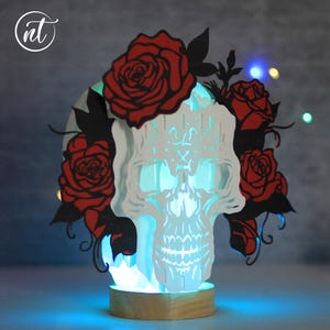 May include: A layered, illuminated skull-shaped decorative piece with red roses and black leaves. The skull is white with intricate cutouts, and the roses are a vibrant red. The base is a light-colored wood, and the piece emits a blue glow.