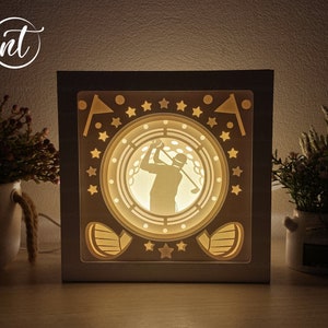 May include: A white wooden shadow box with a golf-themed design. The box features a silhouette of a golfer swinging a club, surrounded by stars and golf flags. The box is illuminated from behind, creating a warm glow.