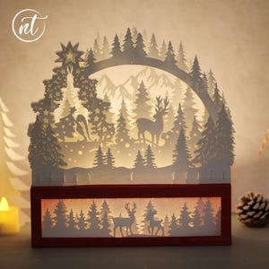 May include: A white paper Christmas scene with a deer and forest design, illuminated by warm light. The scene is set in a dark red wooden frame. A star tops the design, and the scene includes a reindeer, trees, and mountains.