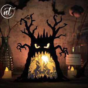 Op de afbeelding: Een zwarte papieren Halloween-boom met een griezelig gezicht en gloeiende ogen. De boom is van binnenuit verlicht, waardoor een sc&egrave;ne met heksen en vleermuizen zichtbaar wordt.
