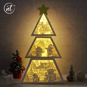 Könnte beinhalten: Eine dreistufige, weiße, dreieckige Weihnachtsbaumdekoration mit warmer Innenbeleuchtung. Jeder Abschnitt zeigt filigrane Scherenschnitt-Silhouetten von Schneemännern, einer Hütte und Bäumen. Die Spitze ist mit einem grünen Stern verziert.