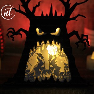 Op de afbeelding: Een zwarte papieren Halloween-boom met een eng gezicht en gloeiende ogen. De boom heeft een uitgesneden ontwerp dat een sc&egrave;ne met heksen en skeletten in de boom laat zien.