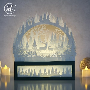 May include: A white paper-cut light box featuring a layered forest scene with a deer and a fawn standing among pine trees. The arched design is set within a rectangular black frame, illuminated from behind to create a glowing, multi-dimensional effect.