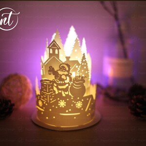 K&ouml;nnte beinhalten: Eine wei&szlig;e Papierlaterne mit einem ausgeschnittenen Design von Weihnachtsmann, der mit einem Sack voller Geschenke einen Schornstein hinaufklettert. Die Laterne wird von innen beleuchtet und erzeugt einen warmen Schein auf die umliegende Schneelandschaft.