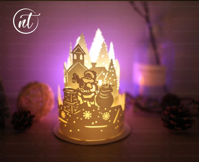 K&ouml;nnte beinhalten: Eine wei&szlig;e Papierlaterne mit einem ausgeschnittenen Design von Weihnachtsmann, der mit einem Sack voller Geschenke einen Schornstein hinaufklettert. Die Laterne wird von innen beleuchtet und erzeugt einen warmen Schein auf die umliegende Schneelandschaft.