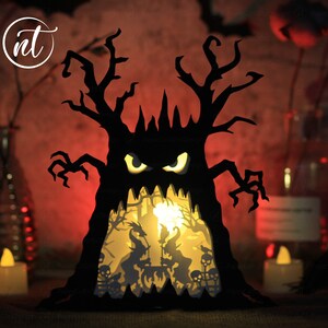 Op de afbeelding: Een zwarte papieren Halloween-boom met een griezelig gezicht en gloeiende ogen. De boom wordt van binnenuit verlicht, waardoor een sc&egrave;ne met heksen en skeletten zichtbaar wordt.