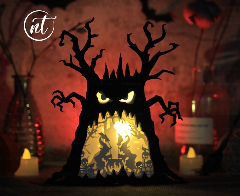 Op de afbeelding: Een zwarte papieren Halloween-boom met een griezelig gezicht en gloeiende ogen. De boom wordt van binnenuit verlicht, waardoor een sc&egrave;ne met heksen en skeletten zichtbaar wordt.