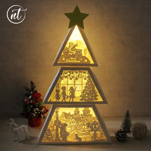 Può includere: Una scatola luminosa in carta bianca ritagliata a tre livelli a forma di albero di Natale. Il livello superiore mostra Babbo Natale con il testo Merry Christmas. Il livello centrale raffigura bambini vicino a un albero. Il basso mostra un villaggio innevato.