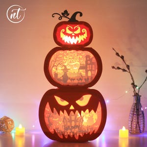 Może przedstawiać: Trzypoziomowa dekoracja na Halloween w kształcie dyni z wyrzeźbionymi twarzami i wewnętrznym oświetleniem. Środkowa warstwa wyświetla napis "Trick or Treat". Dynie świecą, tworząc upiorny efekt. Świece i lampki choinkowe wzmacniają świąteczną scenę.