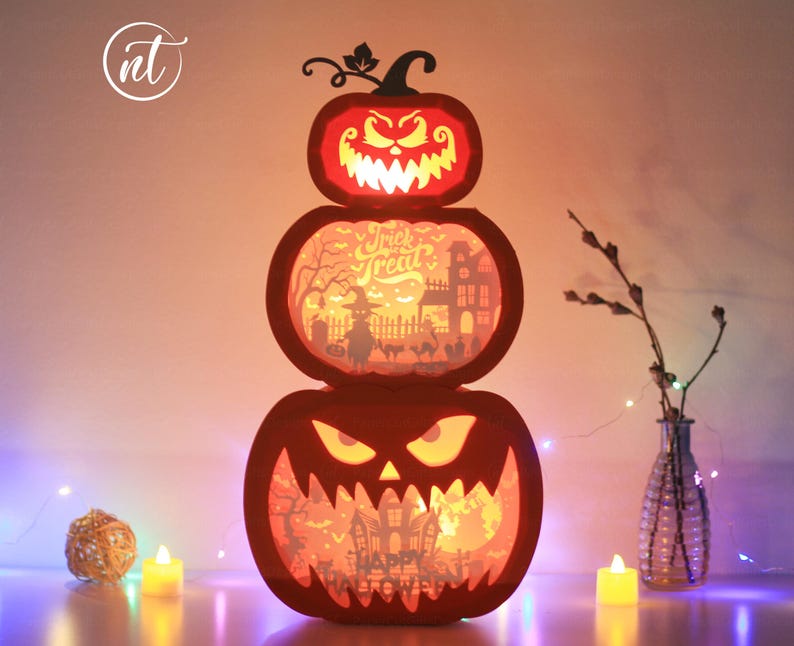 Może przedstawiać: Trzypoziomowa dekoracja na Halloween w kształcie dyni z wyrzeźbionymi twarzami i wewnętrznym oświetleniem. Środkowa warstwa wyświetla napis "Trick or Treat". Dynie świecą, tworząc upiorny efekt. Świece i lampki choinkowe wzmacniają świąteczną scenę.