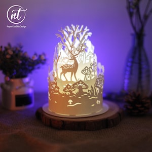 May include: A cylindrical white paper-cut lantern featuring a detailed silhouette of a deer standing among mushrooms and trees. The intricate design is illuminated from within, casting a soft glow. The logo nt PaperCutGiftsDesign is visible.