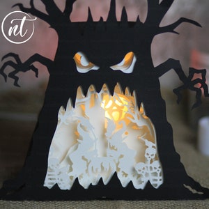 Op de afbeelding: Een zwarte papieren boom met een eng gezicht en gloeiende ogen. De boom wordt van binnenuit verlicht, waardoor een griezelige silhouet ontstaat. De boom is een Halloween-decoratie.