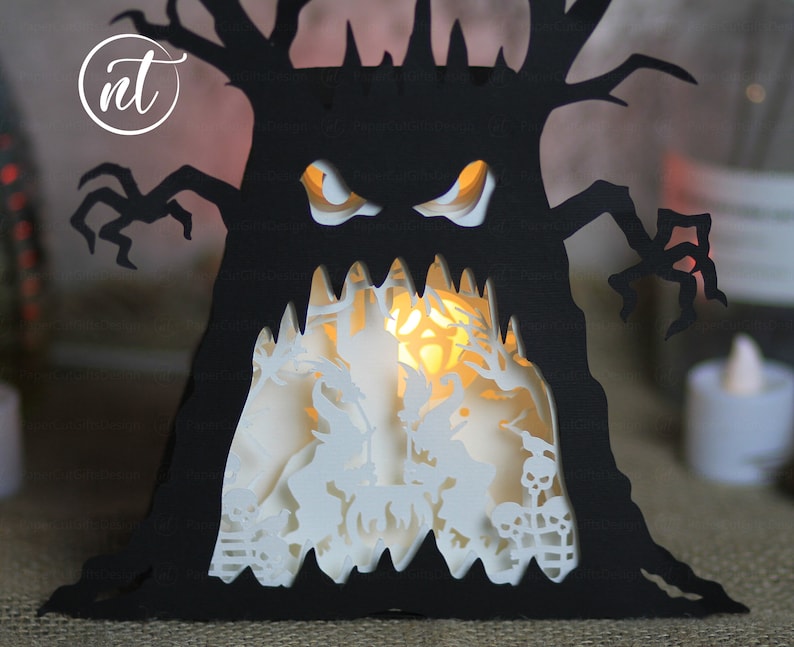 Op de afbeelding: Een zwarte papieren boom met een eng gezicht en gloeiende ogen. De boom wordt van binnenuit verlicht, waardoor een griezelige silhouet ontstaat. De boom is een Halloween-decoratie.