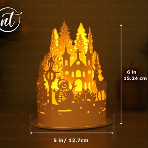 K&ouml;nnte beinhalten: Eine wei&szlig;e Weihnachtsdorf-Szene aus Papier mit einer Kirche, einem Schneemann, einem Rentier und B&auml;umen. Die Szene wird von innen beleuchtet und erzeugt ein warmes Leuchten. Das Dorf ist 15,24 cm hoch und 12,7 cm breit.