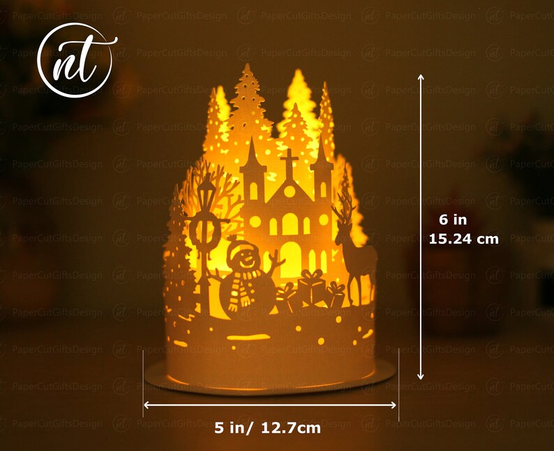 K&ouml;nnte beinhalten: Eine wei&szlig;e Weihnachtsdorf-Szene aus Papier mit einer Kirche, einem Schneemann, einem Rentier und B&auml;umen. Die Szene wird von innen beleuchtet und erzeugt ein warmes Leuchten. Das Dorf ist 15,24 cm hoch und 12,7 cm breit.
