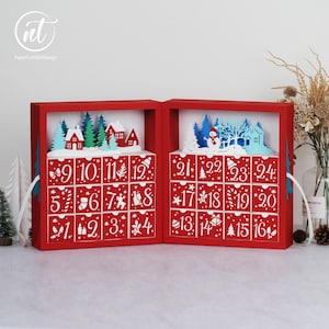 Op de afbeelding: Een rode adventskalender met twee scharnierende delen met genummerde lades van 1 tot 24. Bovenop elk gedeelte staat een gelaagde papieren uitgesneden scène met witte, blauwe en groene bomen, huizen en een sneeuwpop tegen een witte achtergrond.