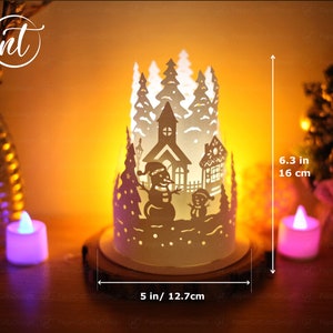 K&ouml;nnte beinhalten: Wei&szlig;er Papierkunst-Kerzenhalter mit einer Winterdorfszene, einschlie&szlig;lich einer Kirche, Schneem&auml;nnern und B&auml;umen. Der Halter ist 16 cm hoch und 12,7 cm breit.
