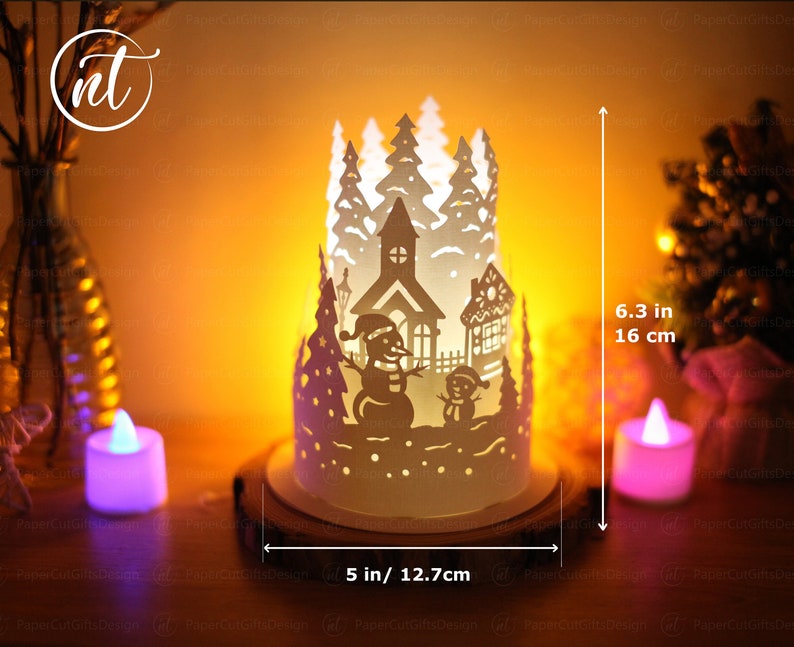 K&ouml;nnte beinhalten: Wei&szlig;er Papierkunst-Kerzenhalter mit einer Winterdorfszene, einschlie&szlig;lich einer Kirche, Schneem&auml;nnern und B&auml;umen. Der Halter ist 16 cm hoch und 12,7 cm breit.