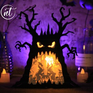 Op de afbeelding: Een zwarte papieren Halloween-boom met een griezelig gezicht en gloeiende ogen. De boom wordt van binnenuit verlicht, waardoor een sc&egrave;ne met heksen en skeletten zichtbaar wordt.