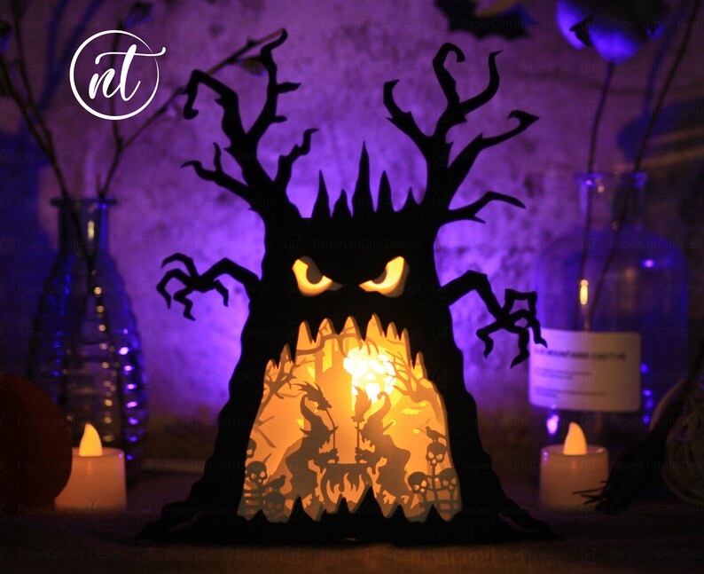 Op de afbeelding: Een zwarte papieren Halloween-boom met een griezelig gezicht en gloeiende ogen. De boom wordt van binnenuit verlicht, waardoor een sc&egrave;ne met heksen en skeletten zichtbaar wordt.