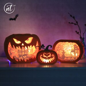 Może przedstawiać: Trzy podświetlane pomarańczowe papierowe dynie z wyrzeźbionymi twarzami i scenami Halloween. Największa dynia ma napis "Happy Halloween". Środkowa dynia jest mniejsza, a prawa dynia ma napis "Trick or Treat". Nad nimi leci sylwetka nietoperza.