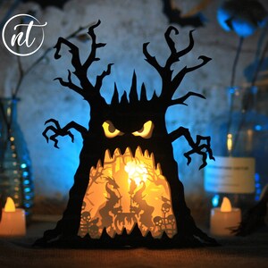 Op de afbeelding: Een zwarte papieren Halloween-boom met een eng gezicht en een gloeiend oranje licht van binnen. De boom is versierd met griezelige silhouetten van heksen, vleermuizen en schedels.