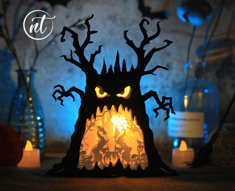 Op de afbeelding: Een zwarte papieren Halloween-boom met een eng gezicht en een gloeiend oranje licht van binnen. De boom is versierd met griezelige silhouetten van heksen, vleermuizen en schedels.