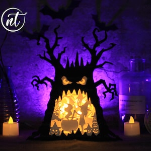 Op de afbeelding: Een zwarte papieren Halloween-boom met een griezelig gezicht en een gloeiend oranje interieur. De boom wordt van binnenuit verlicht met een warm wit licht. De boom is omringd door kleine papieren figuren en een ketel. De boom staat op een tafel met twee brandende kaarsen.