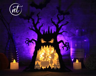 May include: A black papercraft Halloween tree with a scary face and a glowing orange interior. The tree is lit from within with a warm white light. The tree is surrounded by small papercraft figures and a cauldron. The tree is sitting on a table with two lit candles.