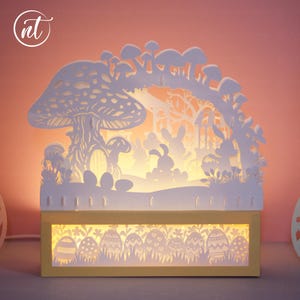 May include: A layered paper Easter scene with a glowing interior. The design features a large mushroom, rabbits, trees, and Easter eggs. The base is a yellow rectangle with egg cutouts, and the scene is illuminated from within.