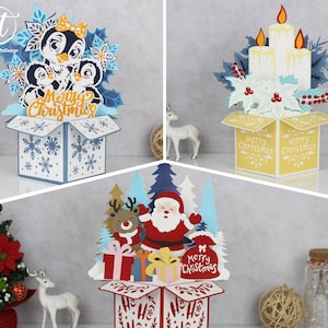 Peut inclure: Trois cartes pop-up sur le thème de Noël. L'une représente des pingouins, une autre des bougies et la dernière le Père Noël avec un renne. Chaque carte contient un message "Joyeux Noël" et des éléments décoratifs.