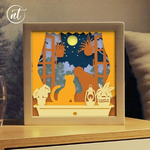 Op de afbeelding: Een 3D-papierkunstwerk met een uitgestanste afbeelding van twee katten die uit een raam kijken naar een nachtscène met een volle maan, sterren en planten. De scène wordt verlicht met een warm geel licht.