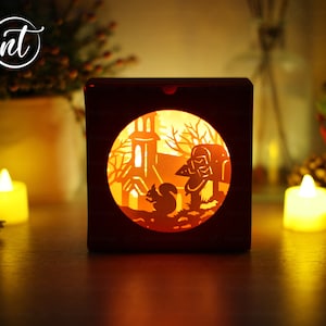 May include: A red wooden box with a cut-out design of a church, a squirrel, and a person with a lantern. The box is lit from inside, creating a warm glow.
