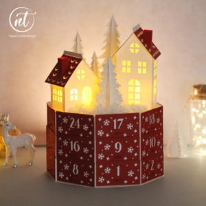 Puede incluir: Un calendario de adviento de papel rojo y blanco con base hexagonal que presenta paneles numerados del 1 al 24 decorados con copos de nieve blancos. La parte superior muestra dos casas de papel blanco iluminadas y varios pinos.