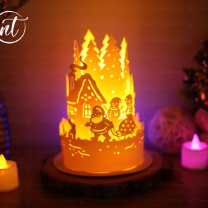 K&ouml;nnte beinhalten: Eine Weihnachts-Papierkunst mit einer brennenden Kerze im Inneren. Die Szene zeigt ein Haus, B&auml;ume, den Weihnachtsmann und Schneem&auml;nner. Die Papierkunst wird von innen beleuchtet und erzeugt ein warmes Leuchten.