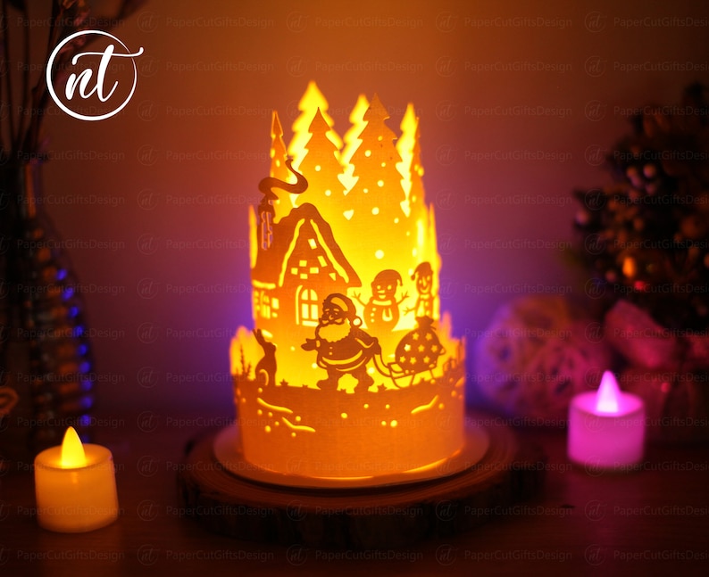 K&ouml;nnte beinhalten: Eine Weihnachts-Papierkunst mit einer brennenden Kerze im Inneren. Die Szene zeigt ein Haus, B&auml;ume, den Weihnachtsmann und Schneem&auml;nner. Die Papierkunst wird von innen beleuchtet und erzeugt ein warmes Leuchten.