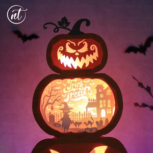 Może przedstawiać: Tr&oacute;jpoziomowa dynia na Halloween z papieru z wyrzeźbioną twarzą i napisem "Trick or Treat". Projekt zawiera nietoperze, wiedźmę, nawiedzony dom i scenę cmentarza, wszystko podświetlone od wewnątrz.