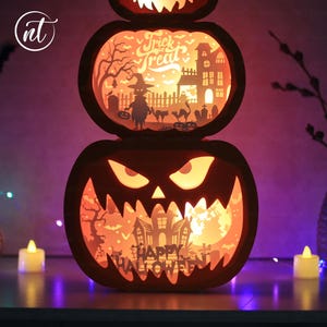 Może przedstawiać: Tr&oacute;jpoziomowa latarnia z dyni na Halloween. G&oacute;rna część zawiera napis "Trick or Treat" ze sceną z wiedźmą i nawiedzonym domem. Środkowa część ma upiorną scenę, a dolna część zawiera napis "Happy Halloween". Latarnia jest podświetlana od wewnątrz.