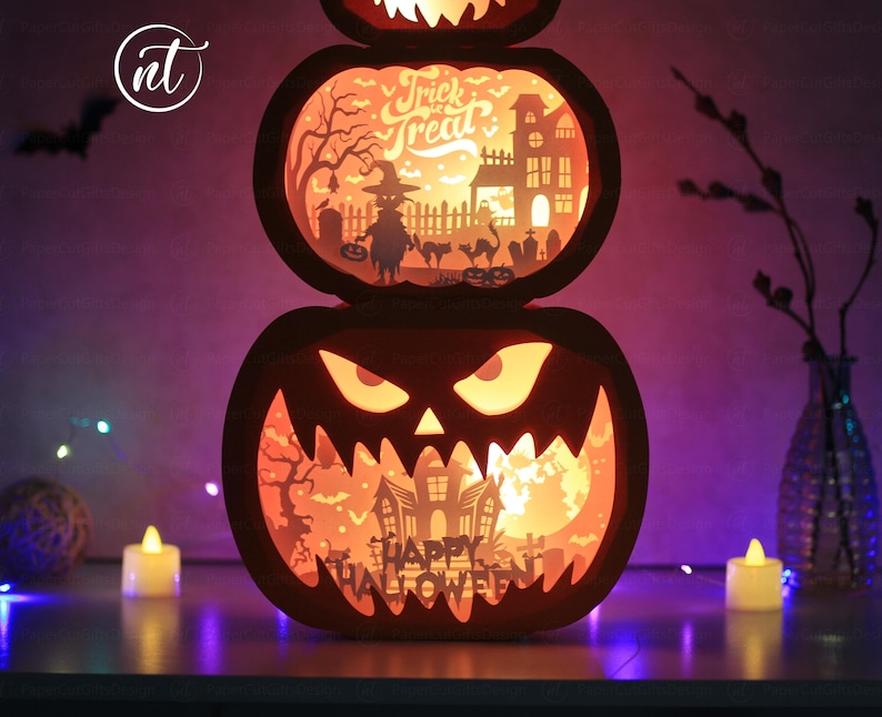 Może przedstawiać: Tr&oacute;jpoziomowa latarnia z dyni na Halloween. G&oacute;rna część zawiera napis "Trick or Treat" ze sceną z wiedźmą i nawiedzonym domem. Środkowa część ma upiorną scenę, a dolna część zawiera napis "Happy Halloween". Latarnia jest podświetlana od wewnątrz.