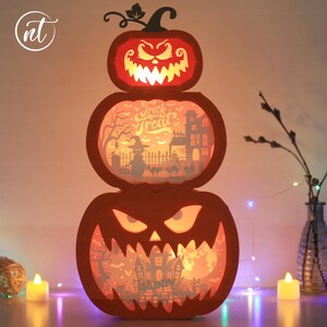 Może przedstawiać: Trzypoziomowa dekoracja na Halloween w kształcie dyni. G&oacute;rna dynia ma wyrzeźbioną twarz z groźnym wyrazem. Środkowa dynia ma napis "Trick or Treat" i upiorną scenę. Dolna dynia ma wyrzeźbioną twarz.