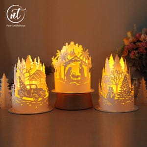 May include: Three white paper-cut cylindrical lanterns illuminated from within by warm yellow light. The left lantern features a truck and house, the center shows a nativity scene, and the right depicts a snowman, house, and trees.